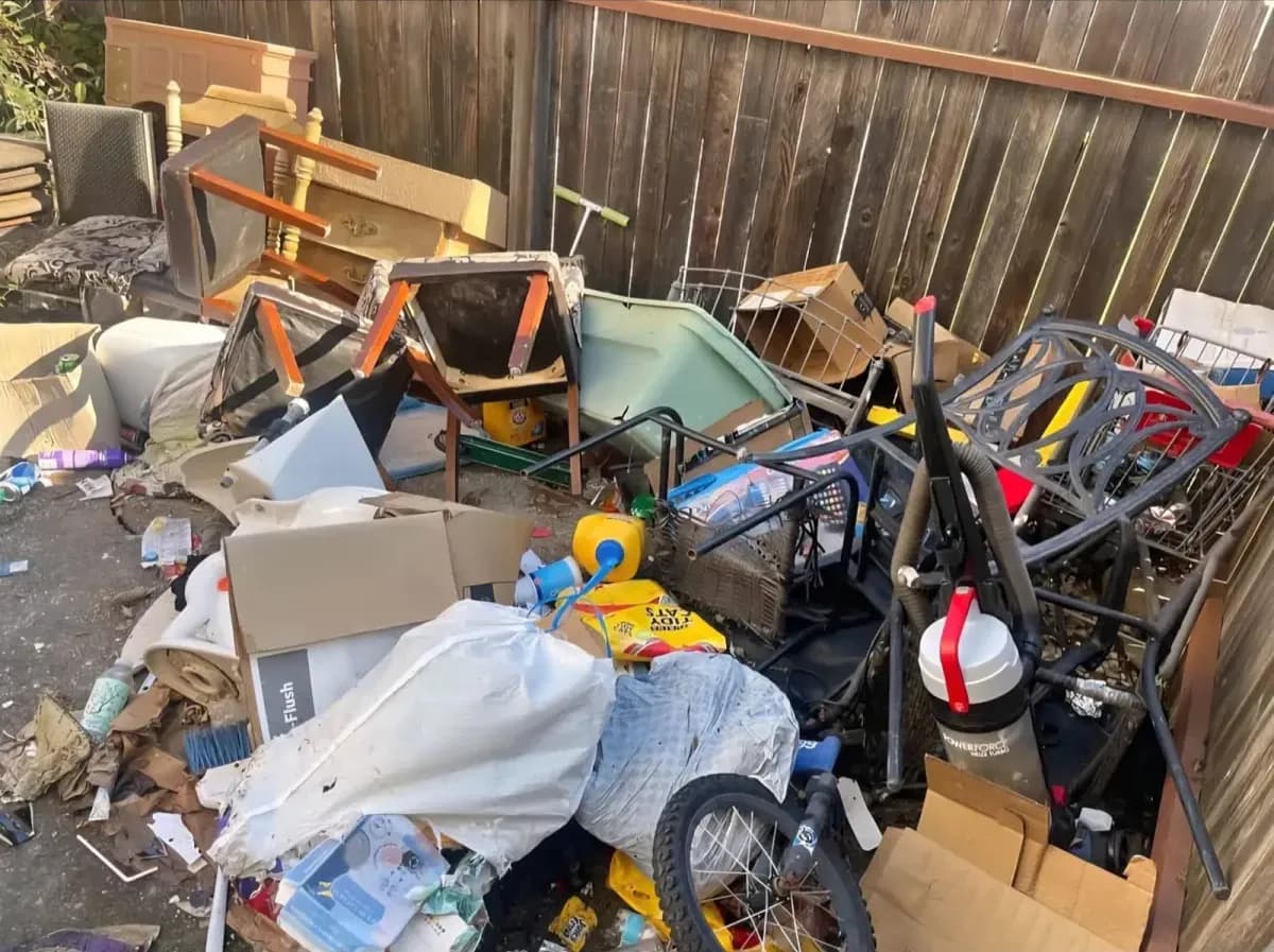 Large backyard pile of furniture, bikes, boxes and bags against a wood fence