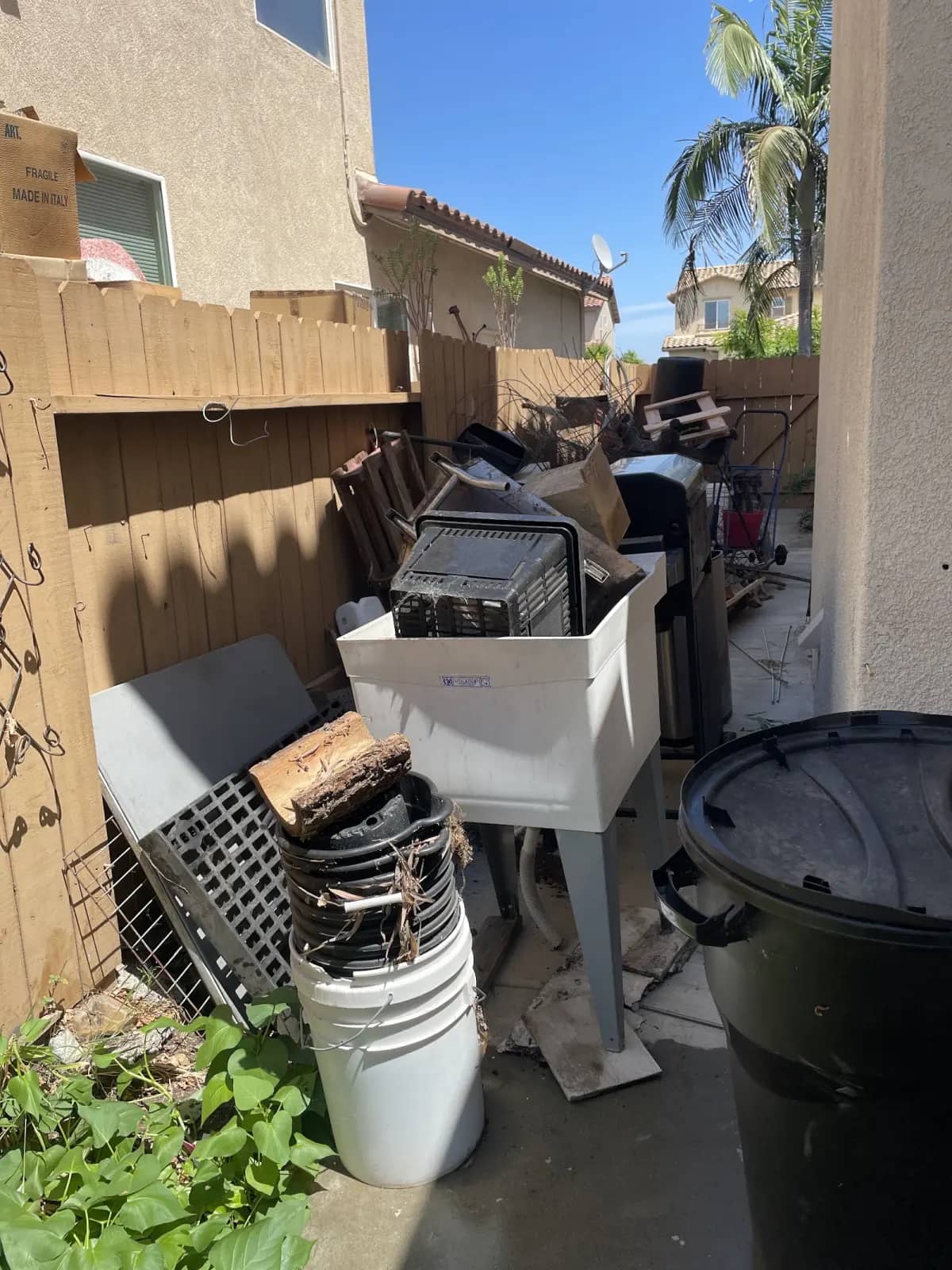 Side yard packed with a utility sink, crates, buckets and trash bins before pickup